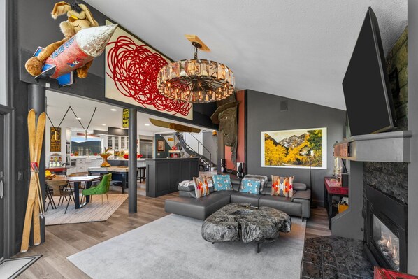 A modern living room with a fireplace, a large sofa, and a dining area with a kitchen in the background.