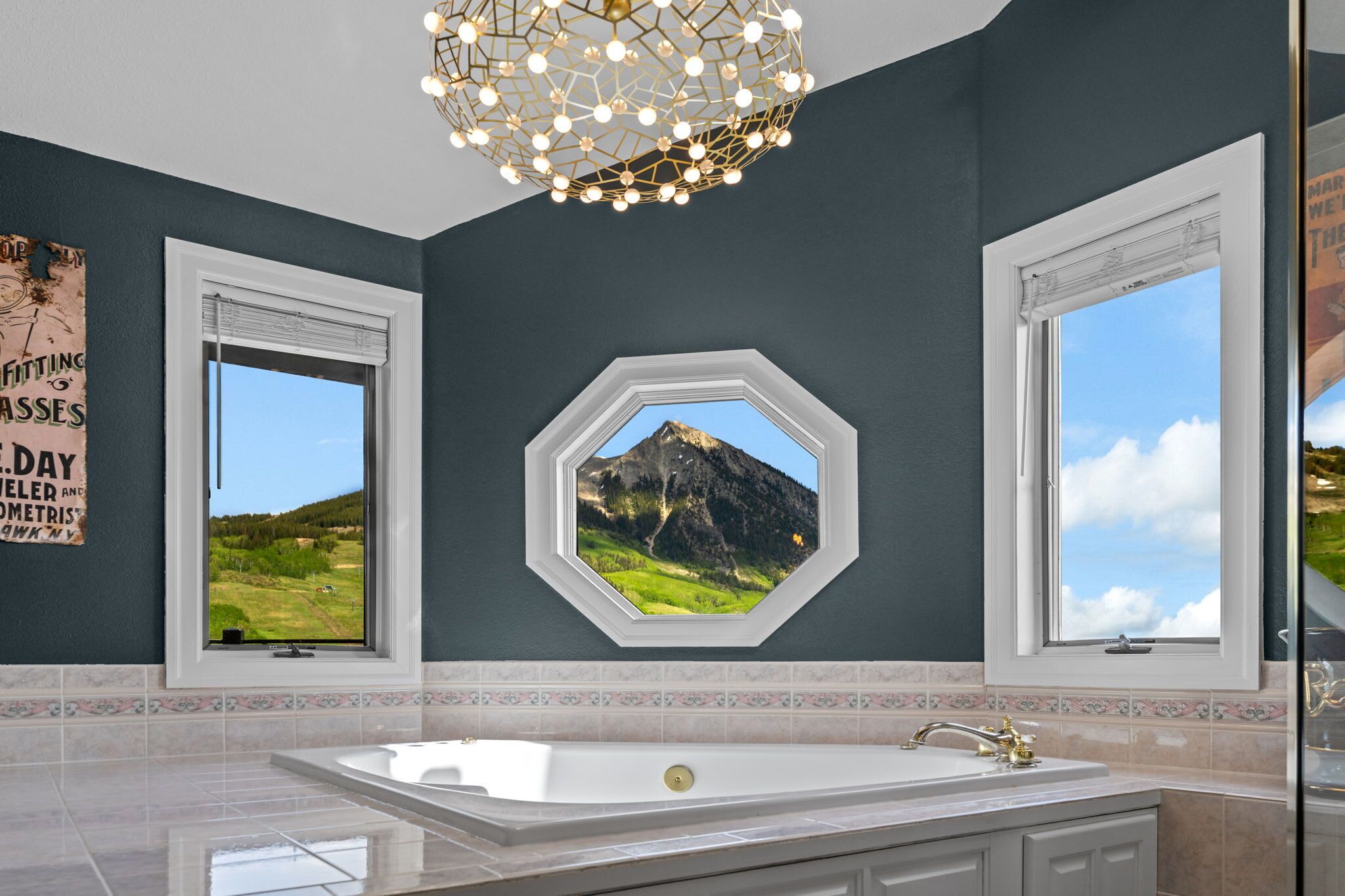 A bathroom with a large bathtub, two windows, a decorative light fixture, and a wall-mounted picture of a mountain landscape.
