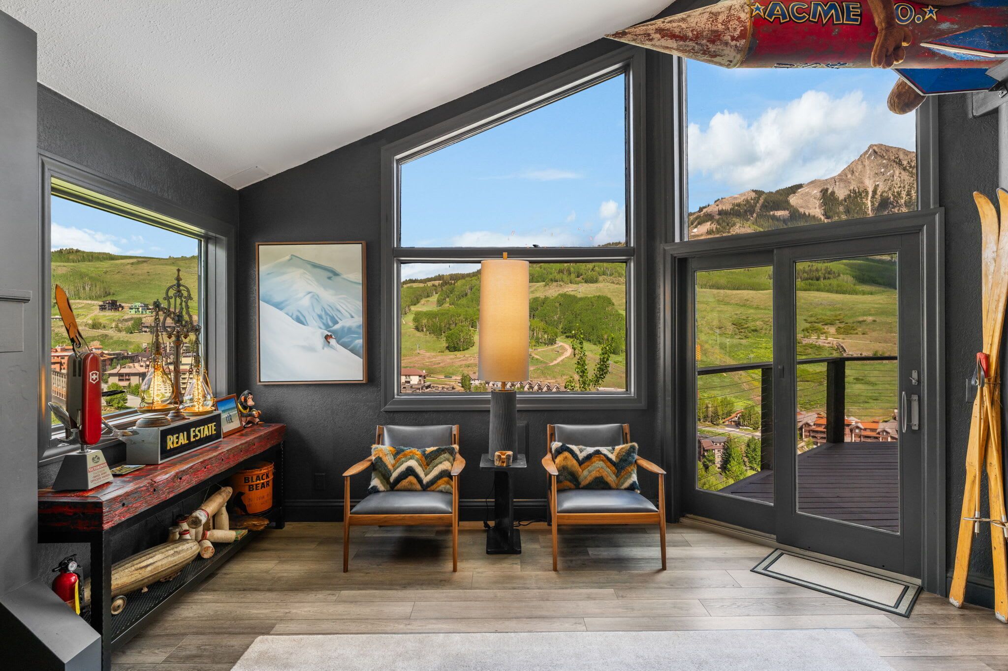 A room with large windows, a fireplace, two chairs, a shelf with items, and a view of a mountainous landscape.