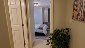Un couloir menant à une chambre à coucher, où se trouvent un lit, un luminaire au plafond et une plante en pot.