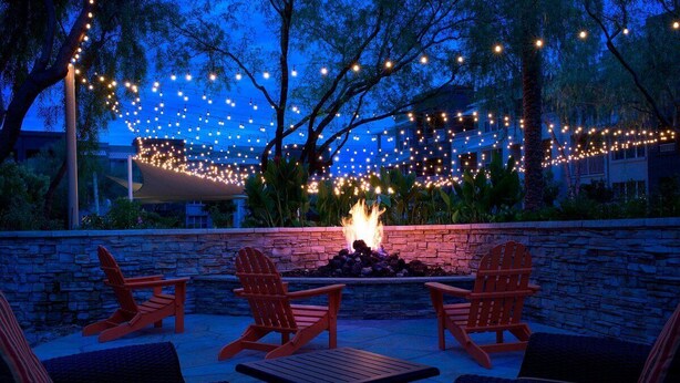 Un espace extérieur aménagé avec des chaises Adirondack et un foyer, le tout entouré de guirlandes lumineuses et d’arbres.