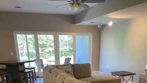 A spacious living room with a large beige sectional sofa, a small wooden coffee table, and a dark wooden desk with a chair. There are large windows providing natural light and a view of greenery outside. - The Spacious Canal Condo (Caseville)
