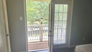 Ein Zimmer mit Holzboden, einem Bett mit weißer Bettdecke, einer Tür zu einem Balkon mit Geländer und einem Fenster mit Jalousien.