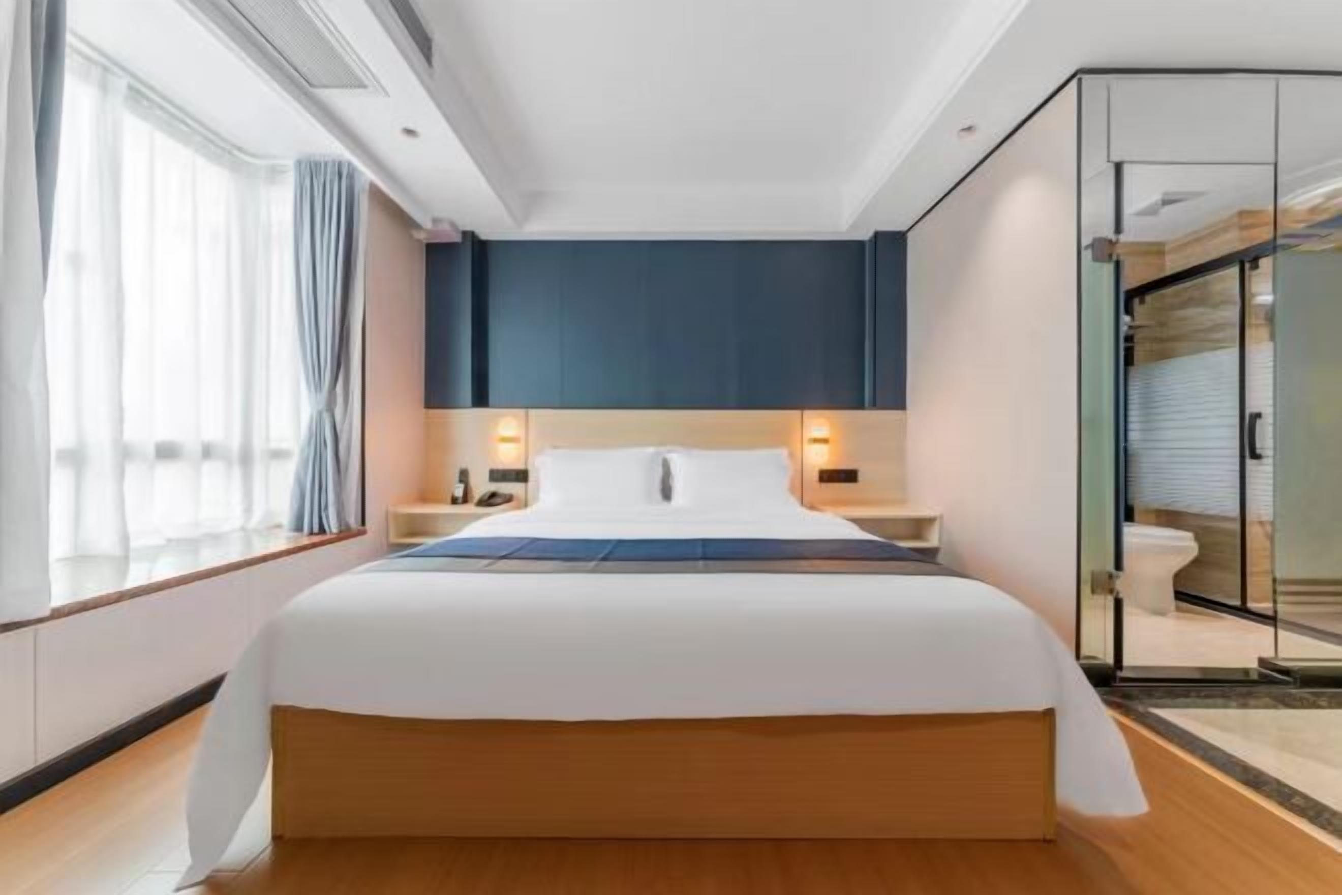 A modern hotel room with a large bed, bedside tables with lamps, a glass shower enclosure, and a view of the city.