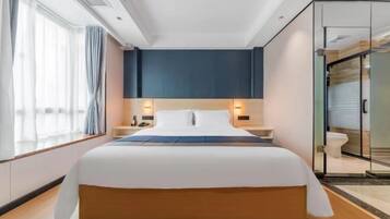 A modern hotel room with a large bed, bedside tables with lamps, a glass shower enclosure, and a view of the city.