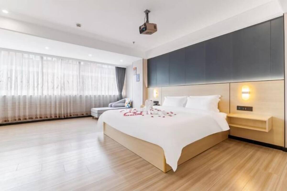 Ein modernes Hotelzimmer mit einem großen Bett, einem Nachttisch, einem an der Wand befestigten Fernseher und einem großen Fenster mit transparenten Vorhängen.