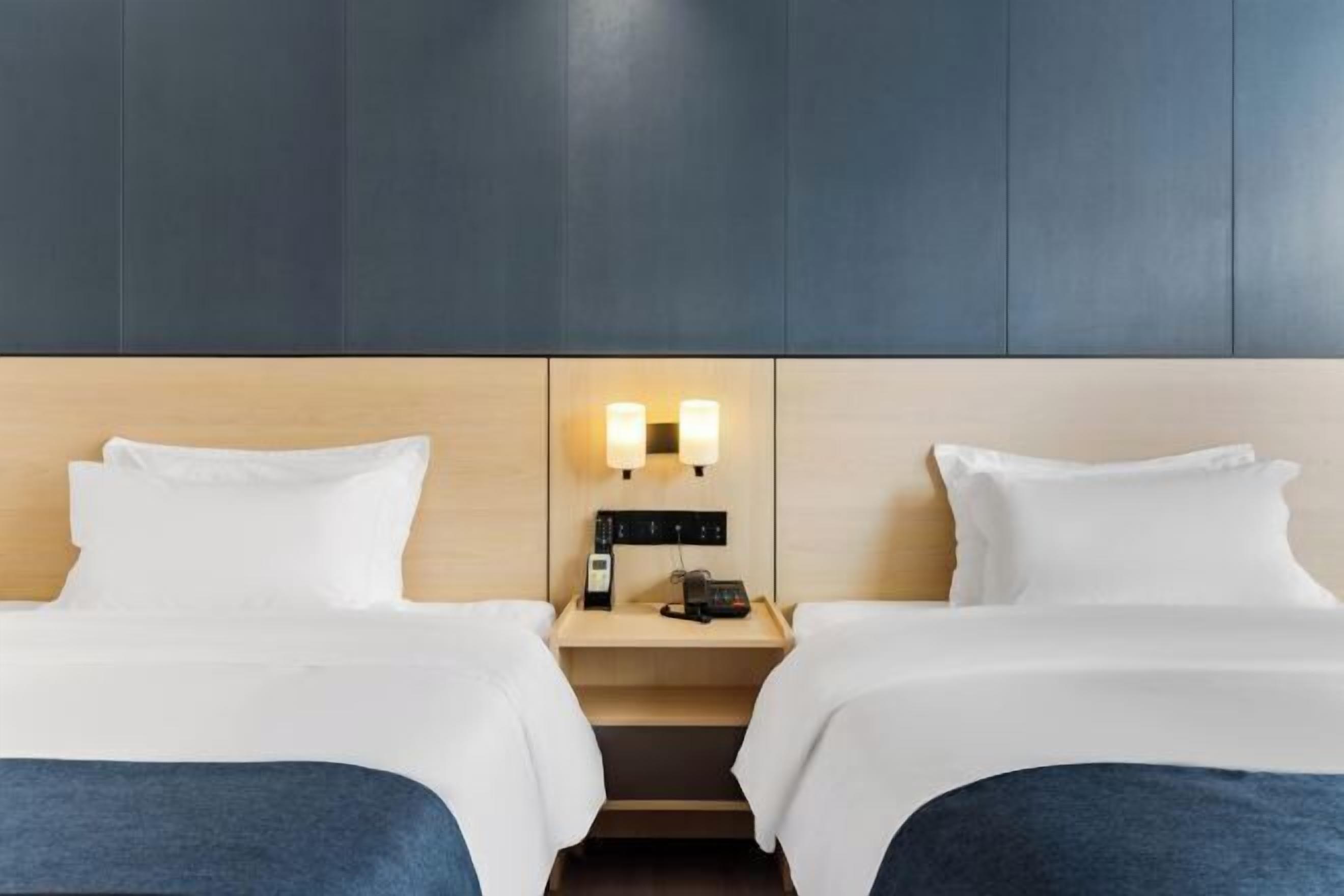 A hotel room with two beds, a nightstand with a phone and lamp, and a wall-mounted light fixture.