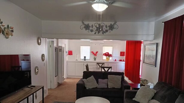 A living room with a sofa, coffee table, and TV stand. There is a kitchen in the background with red curtains.