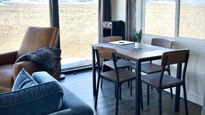 Un intérieur moderne avec un plafond en bois, de grandes fenêtres offrant une vue sur un paysage enneigé, un coin repas avec une table et des chaises, et un canapé en cuir agrémenté de coussins.