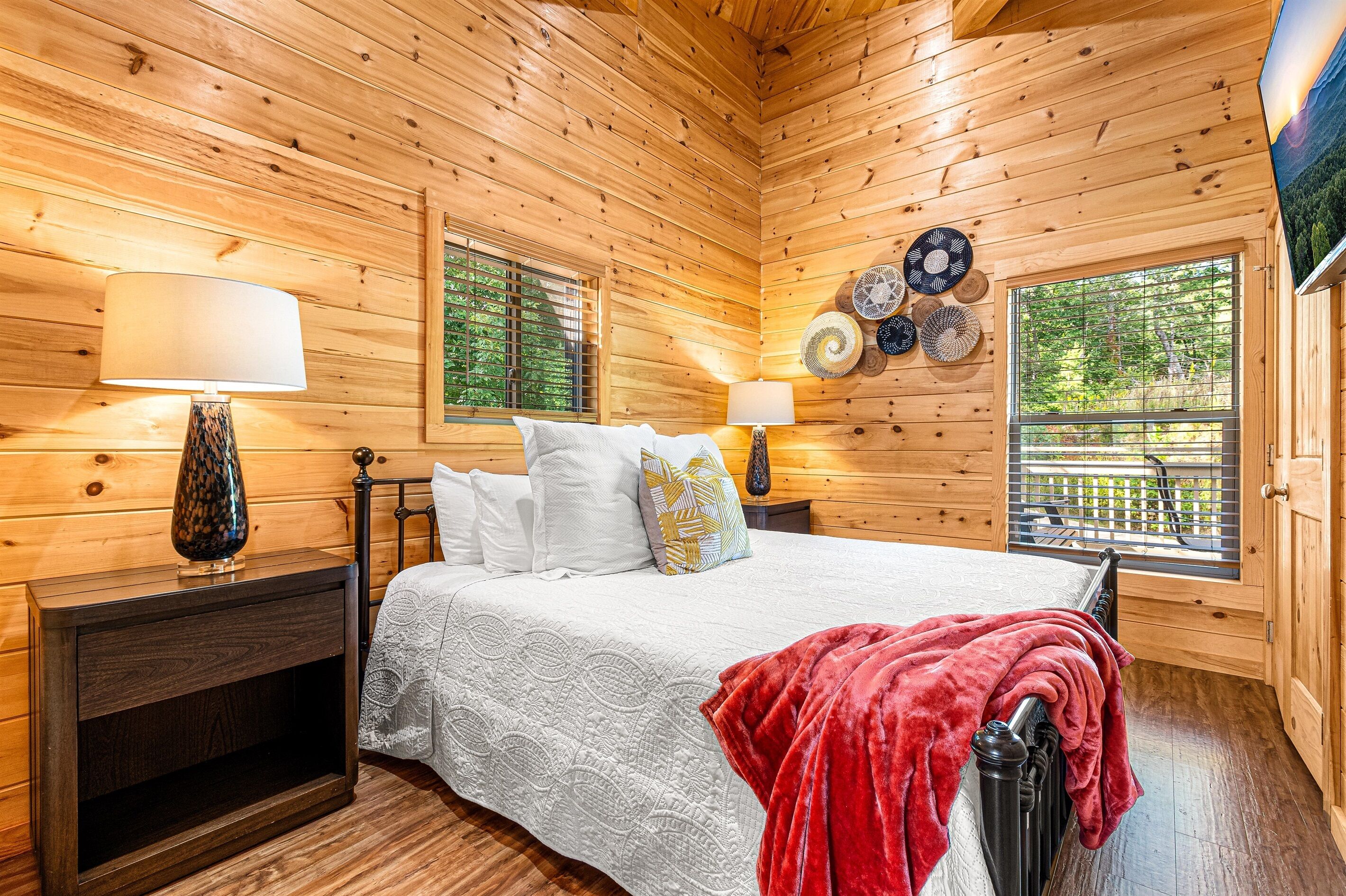 A wooden cabin bedroom with a bed, two bedside tables, a lamp, and a window with blinds.
