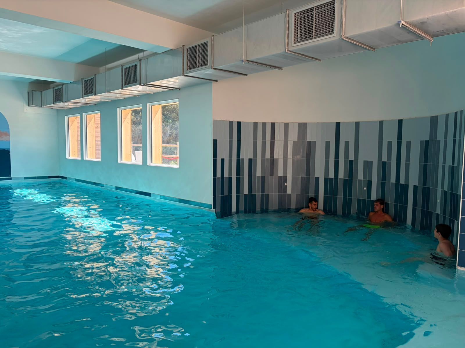 An indoor swimming pool with clear water, large windows, and a tiled wall.