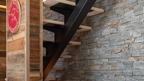 A cozy room with a wooden accent wall, a stone wall, a brown sofa with white pillows, and a staircase with black railings. - Olvera 501 - New apartment on the slopes (VALMEINIER)