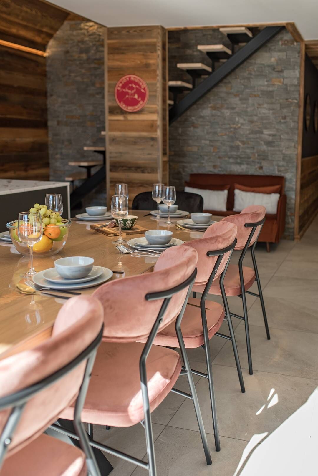 A dining area with a long wooden table set for six, pink upholstered chairs, and a stone wall with a wooden staircase.