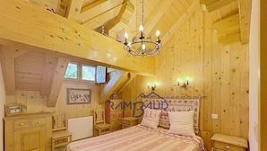 A cozy bedroom with a wooden ceiling, a bed with patterned bedding, a nightstand, a dresser, and a chandelier.