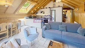 A cozy log cabin interior with a dining area, a living room with a sofa and armchairs, a kitchen with a refrigerator, and a skylight.