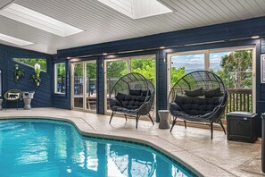An indoor swimming pool with black wicker chairs, a view of the outdoors, and a wooden ceiling.