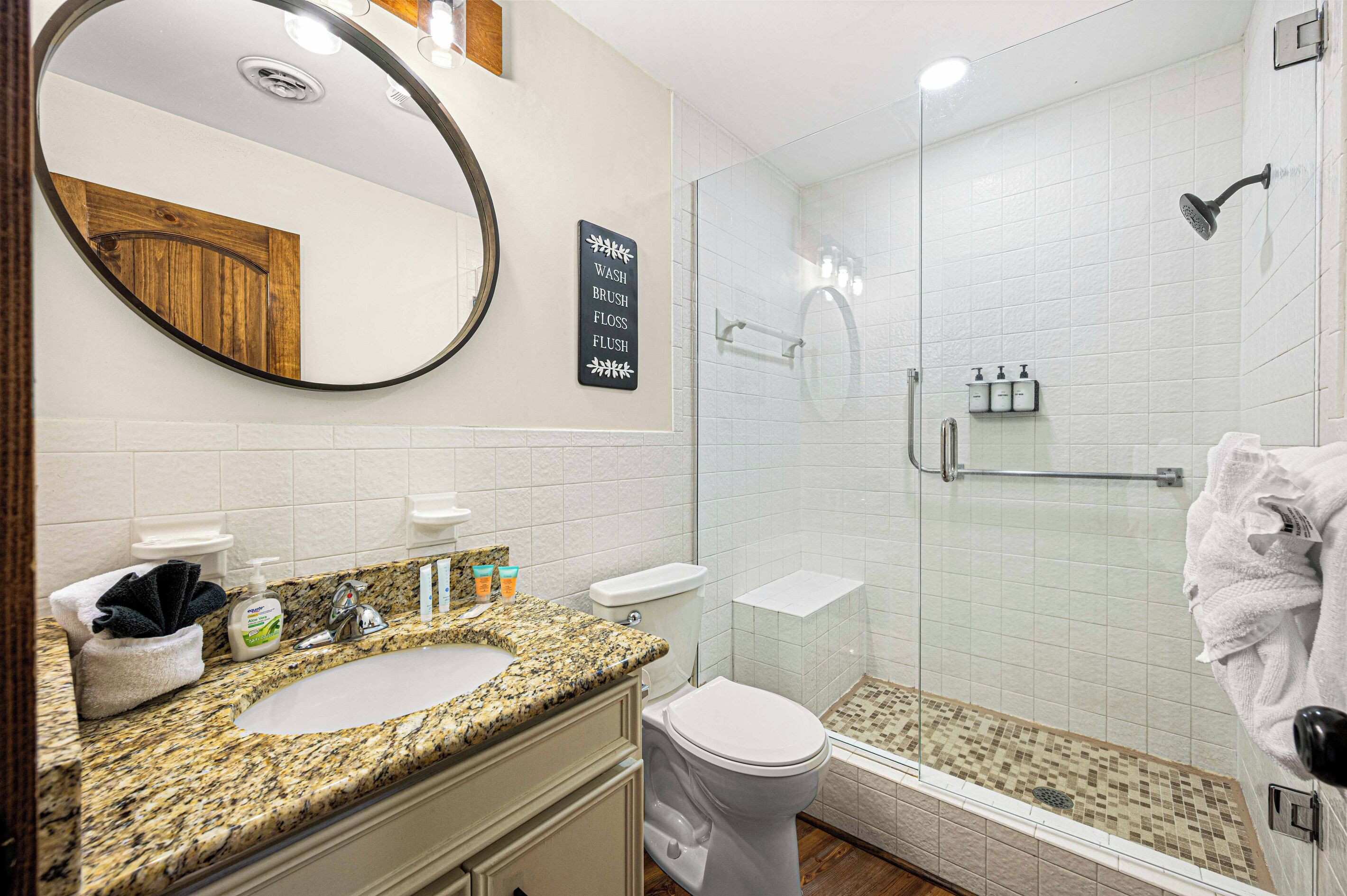 Un baño con encimera de granito, un inodoro, ducha con puerta de cristal y un espejo.