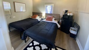A hotel room with two beds, a desk, a chair, a radiator, a window with curtains, and a wall-mounted air conditioning unit.