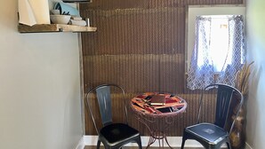 A room with a textured wall, a small round table with chairs, a window with patterned curtains, and a shelf with various items.