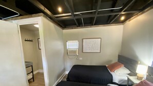 A hotel room with two beds, a small table, a chair, a wall-mounted air conditioner, and a door leading to another room.