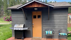 A small wooden cabin with a grill, two chairs with striped cushions, and a door.