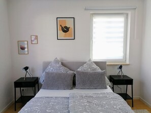 A bedroom with a bed, two nightstands, a window with blinds, and two framed pictures on the wall.