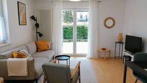 Un salón moderno con un sofá gris, una silla de madera, una mesita, un televisor y un ventanal grande con cortinas.