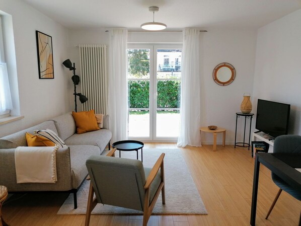 A modern living room with a grey sofa, a wooden chair, a small table, a TV, and a large window with curtains.