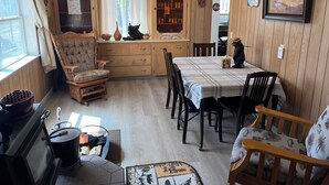 Une pièce chaleureuse comprenant une table à manger, des chaises, un fauteuil en bois, un poêle, une bouilloire et une bibliothèque.