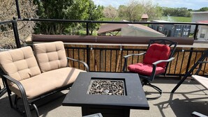 Une terrasse extérieure avec un canapé beige, une chaise rouge et une table carrée dotée d’un foyer.