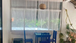 Una habitación con una mesa, sillas, un ventilador de techo, una ventana con cortinas traslúcidas y una planta en maceta.