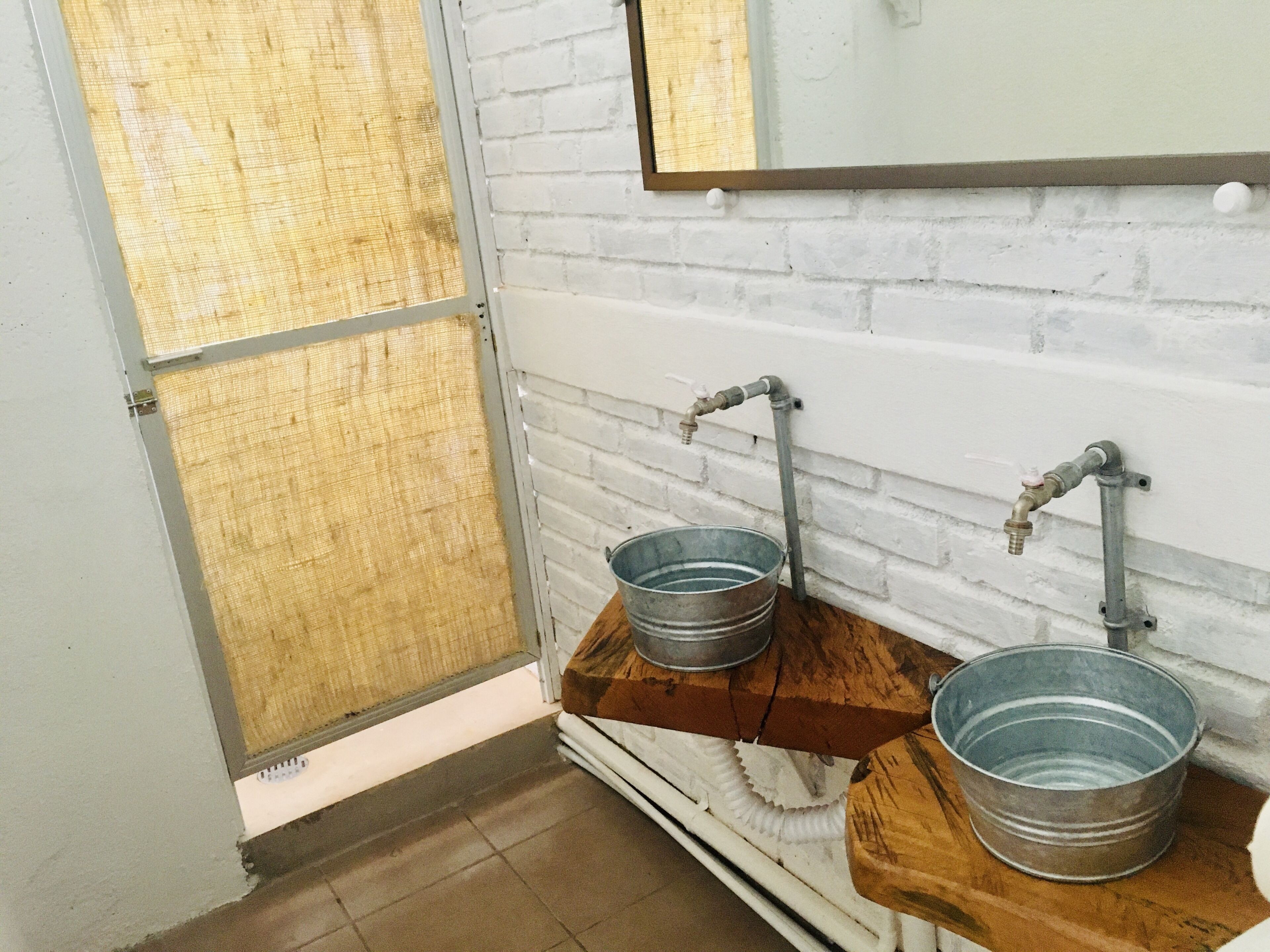 Un bagno con due secchi di metallo su mensole di legno, una parete in mattoni bianchi e uno specchio sopra i lavandini.