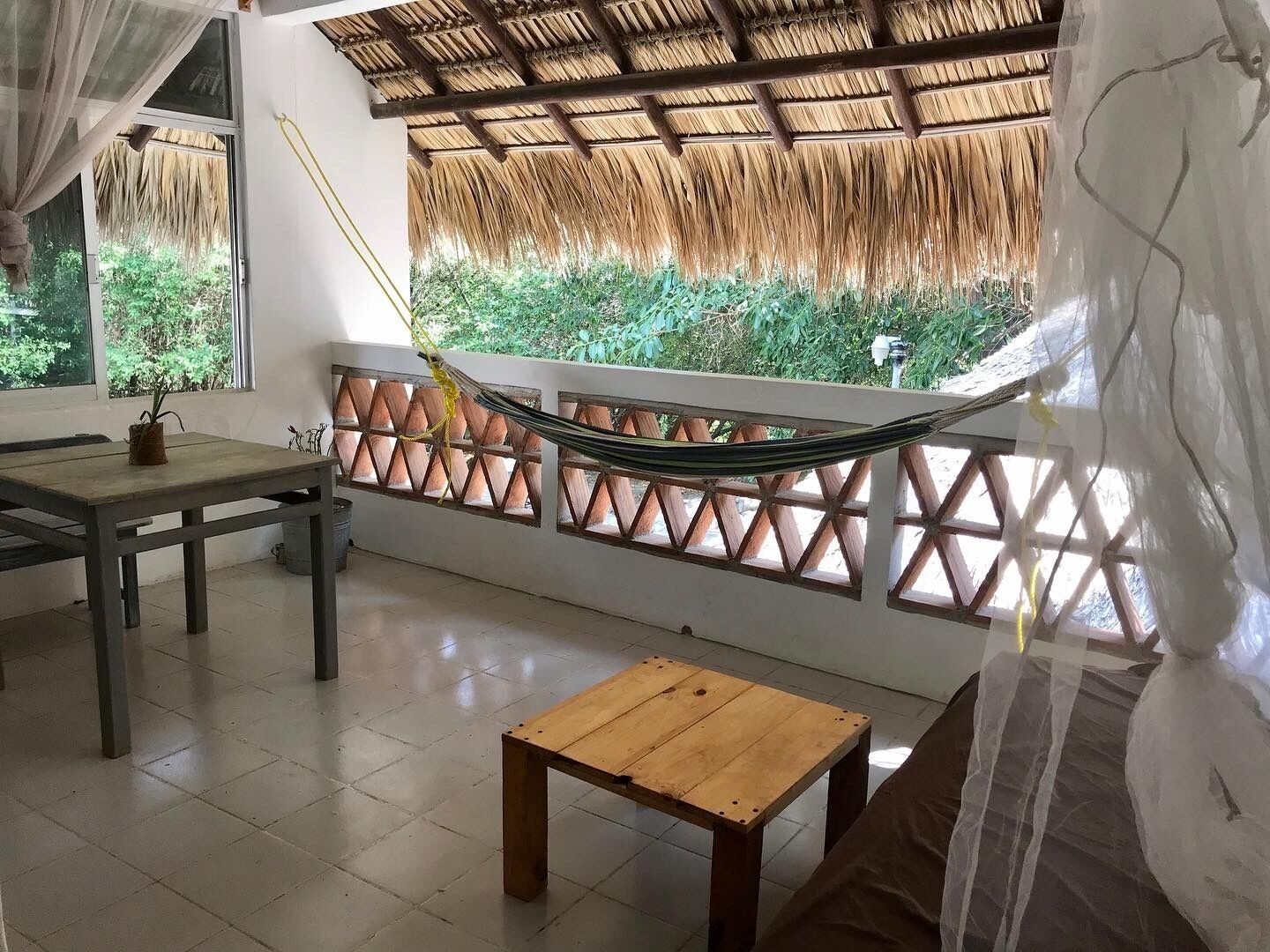 Una habitación con una hamaca, una mesa de madera y vistas a la vegetación a través de una ventana con celosía.
