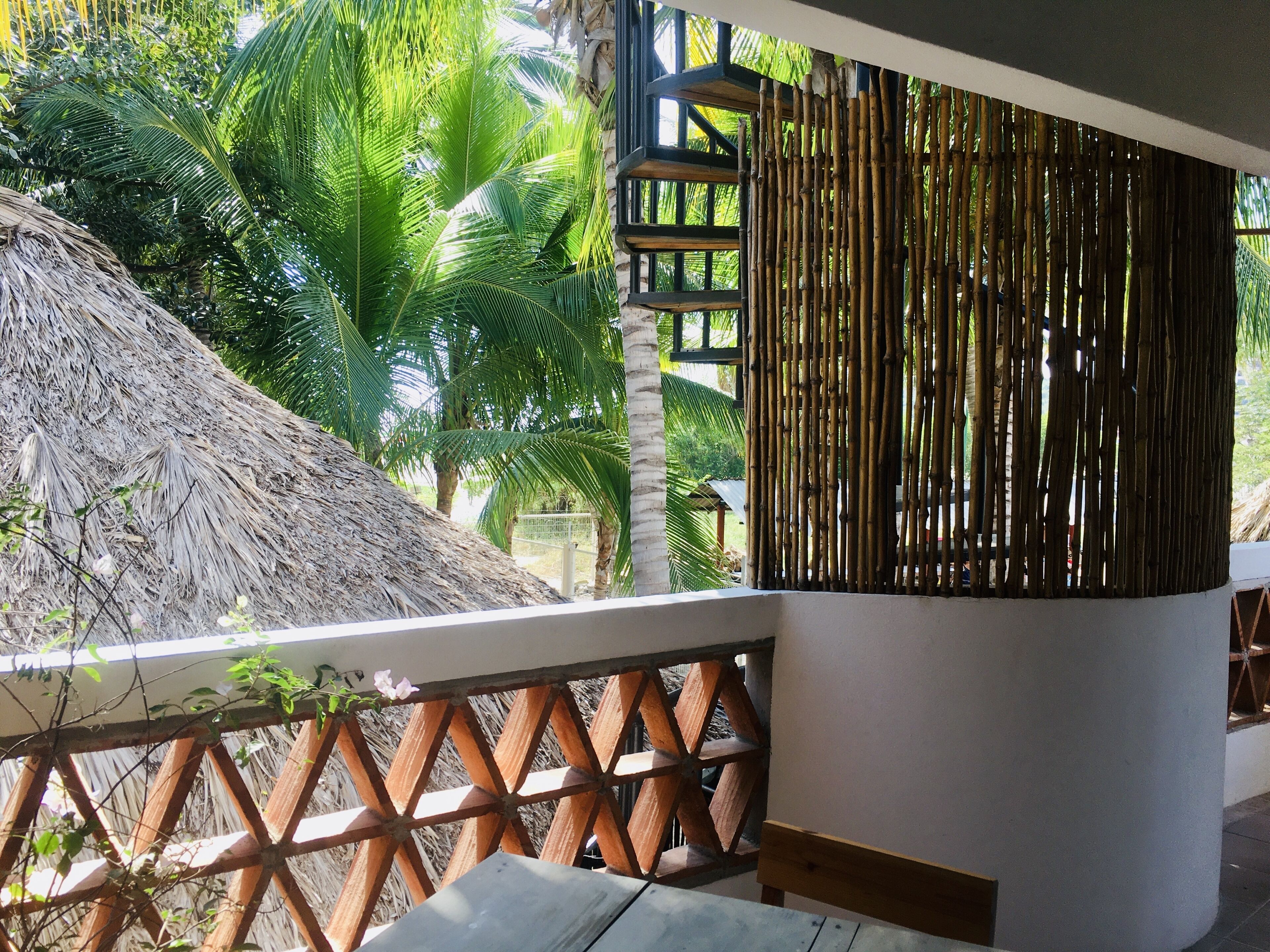 A balcony with a thatched roof, bamboo wall, and a view of palm trees.