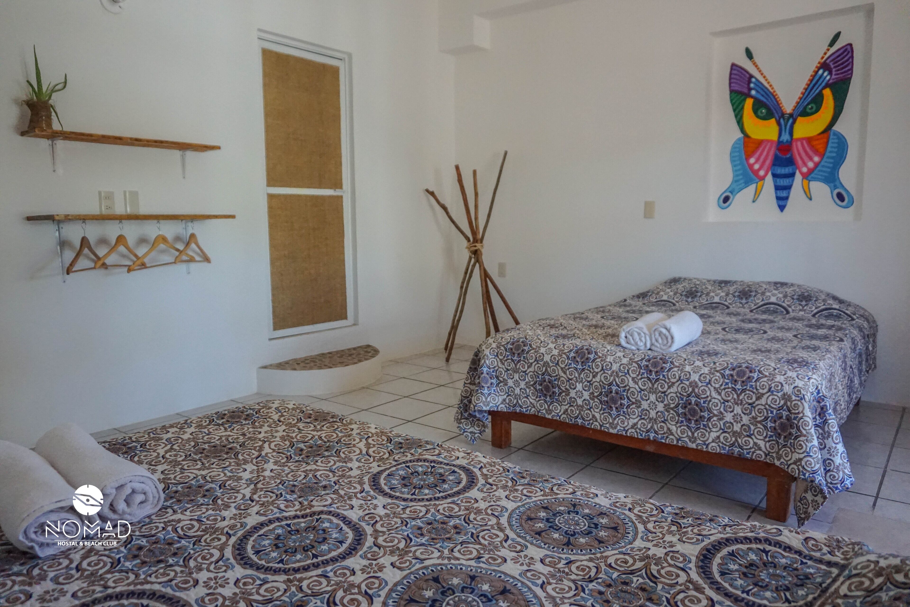 A hotel room with a bed, a patterned rug, a wall-mounted shelf with a plant, a painting, and a towel on the bed.