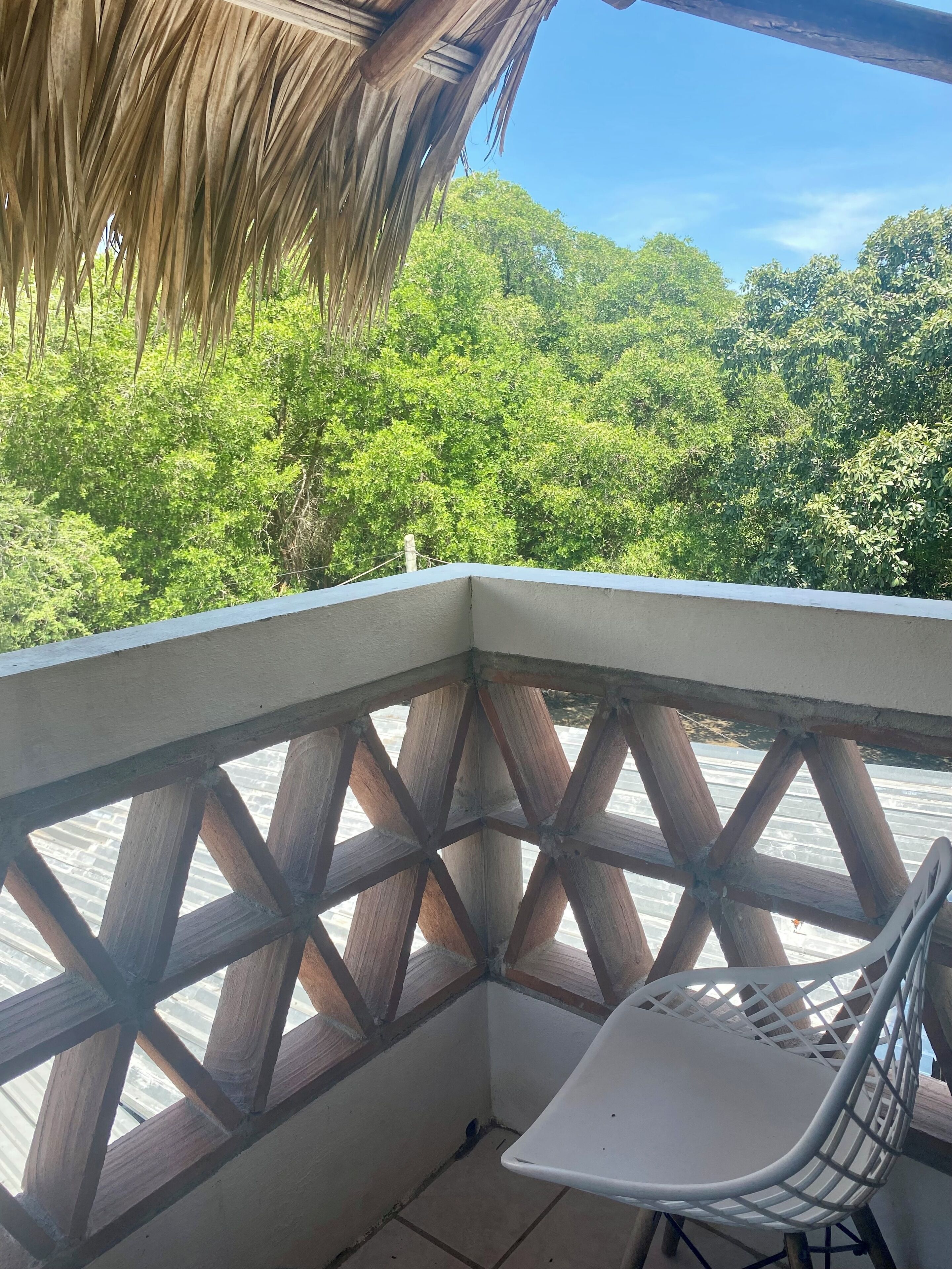 A balcony with a wooden lattice railing, a white chair, and a view of dense green foliage.