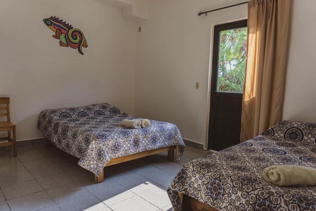 Une chambre avec deux lits, une chaise, un motif décoratif coloré sur le mur et une fenêtre avec des rideaux.