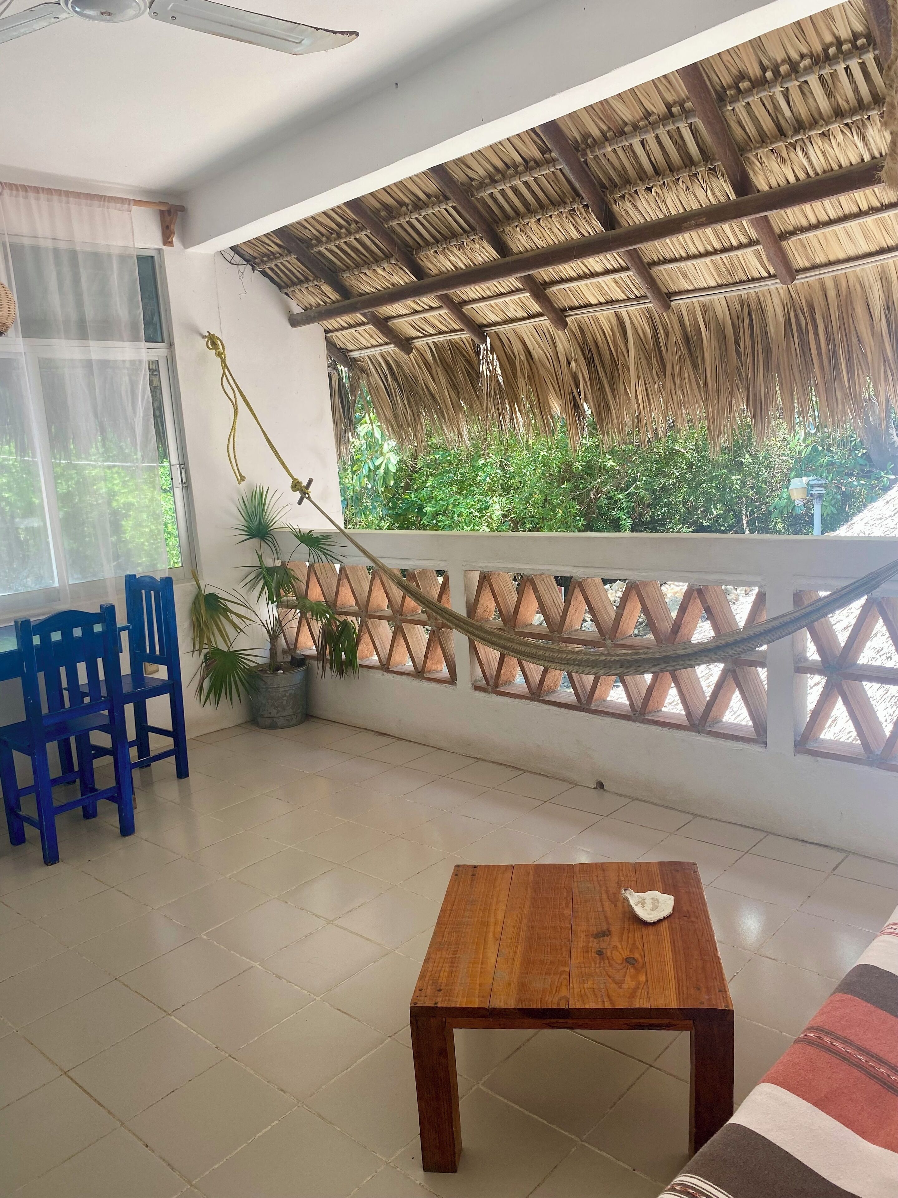 A room with a wooden table, blue chairs, a hammock, and a view of greenery through a window.