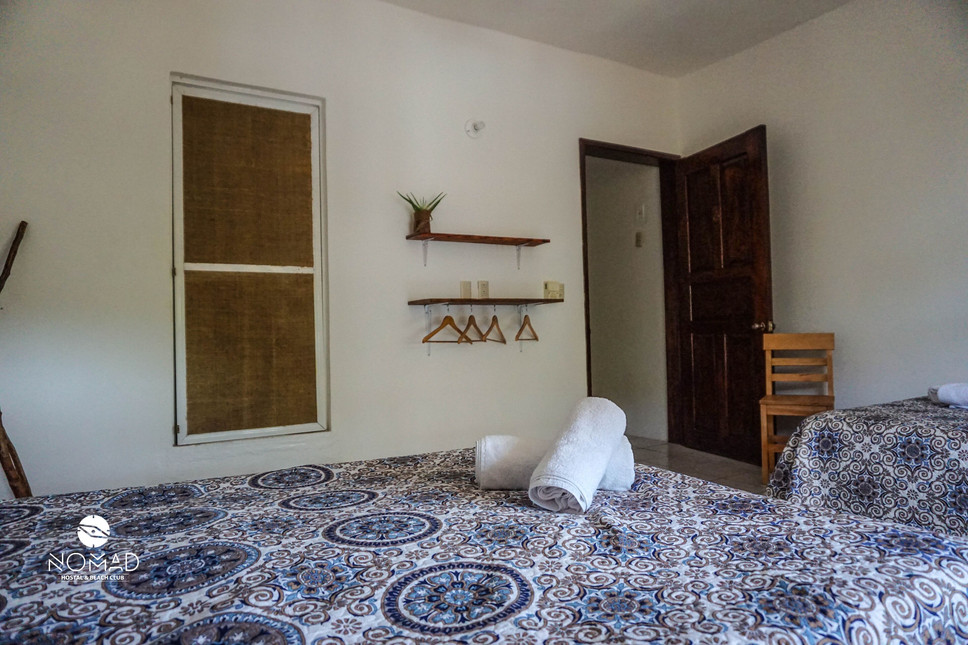 Une chambre d  ’hôtel avec une couvre-lit à motifs, une porte en bois, une étagère ornée d’objets décoratifs et une fenêtre avec un store marron.