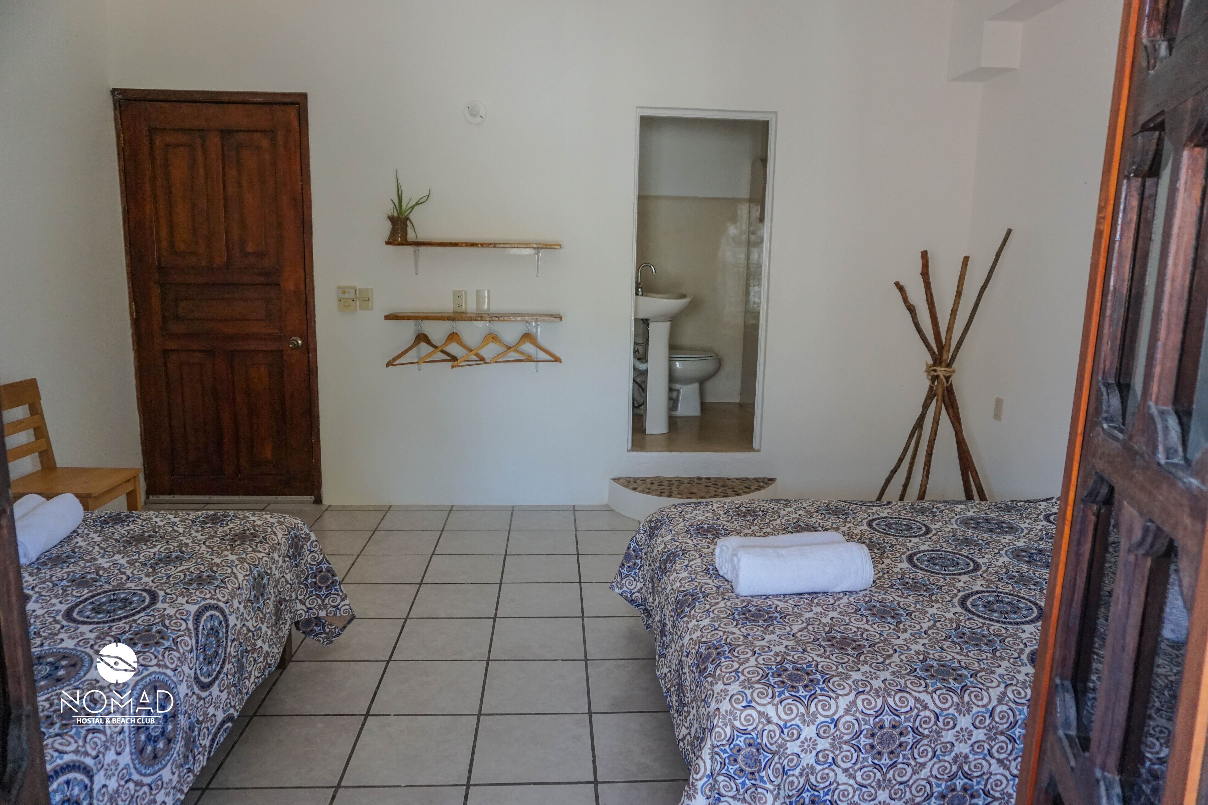 Habitación de hotel con dos camas, una puerta de madera, un baño con lavamanos e inodoro, y un estante pequeño con plantas.