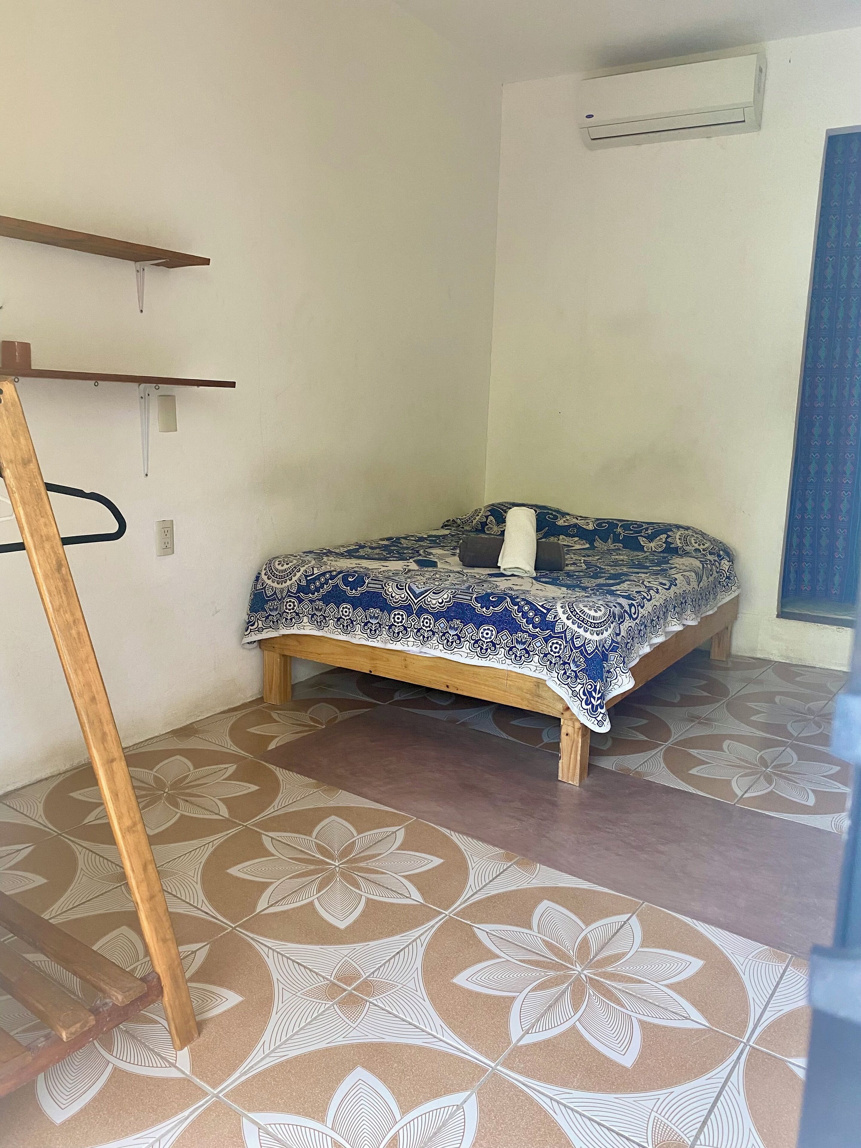A small, simple room with a bed, a wooden bench, and patterned floor tiles.