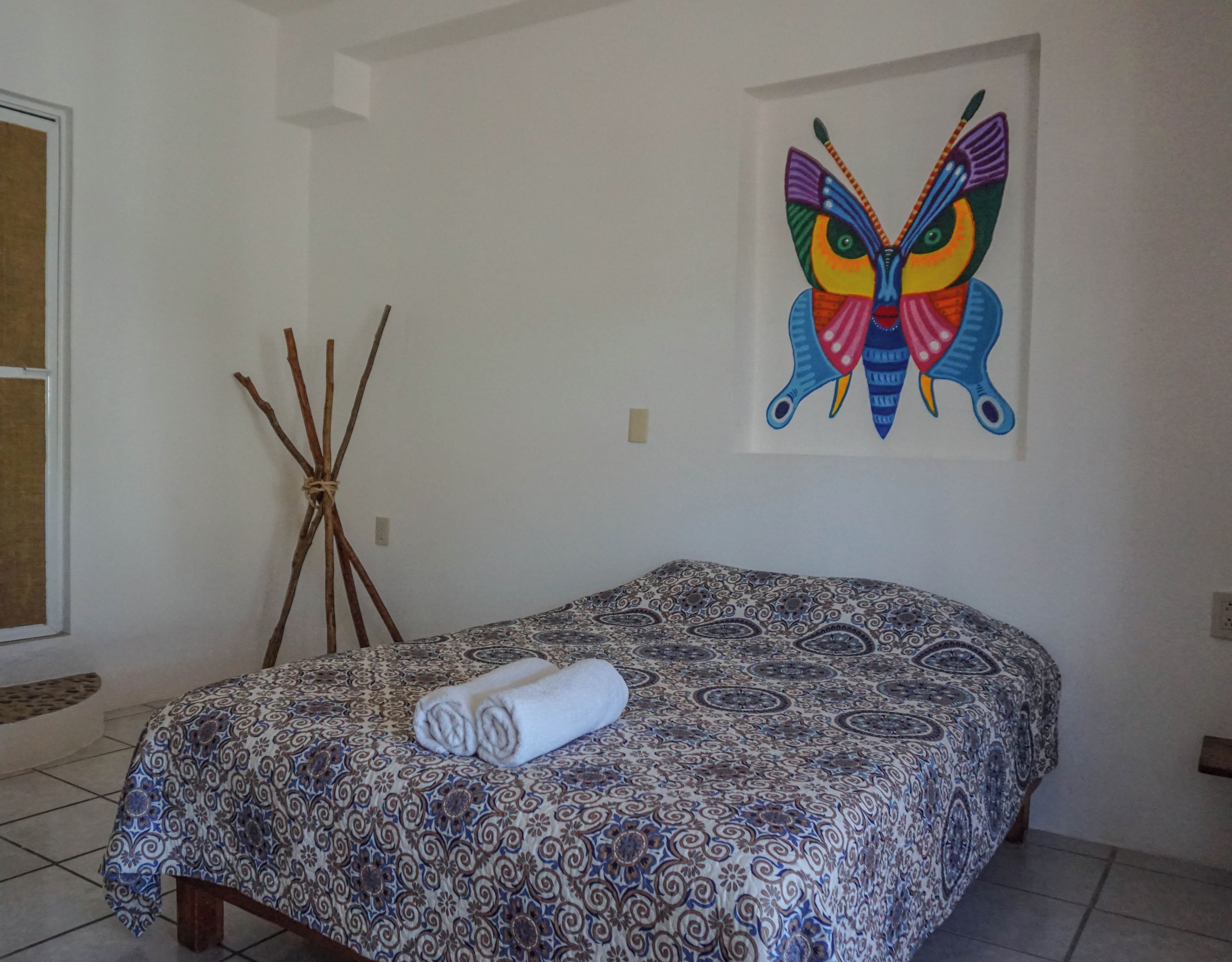 A bedroom with a patterned bedspread, a rolled towel, a tripod, and a large butterfly painting on the wall.