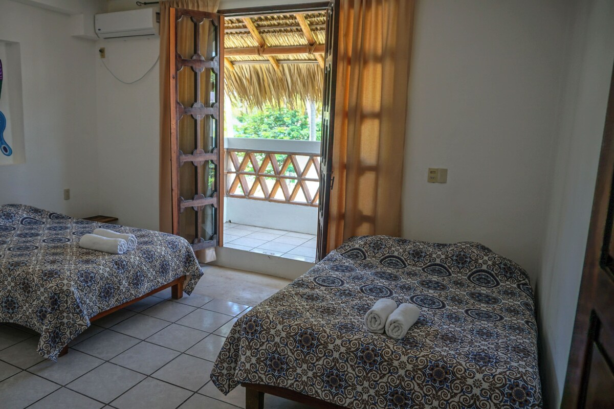 Habitación de hotel con dos camas, una obra de arte de mariposa de colores en la pared y vista al exterior a través de una puerta con techo de paja.