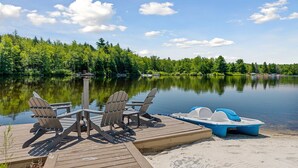 Une jetée en bois, agrémentée de chaises Adirondack et d’un petit bateau, offre une vue sur un lac paisible, avec une forêt dense en arrière-plan.