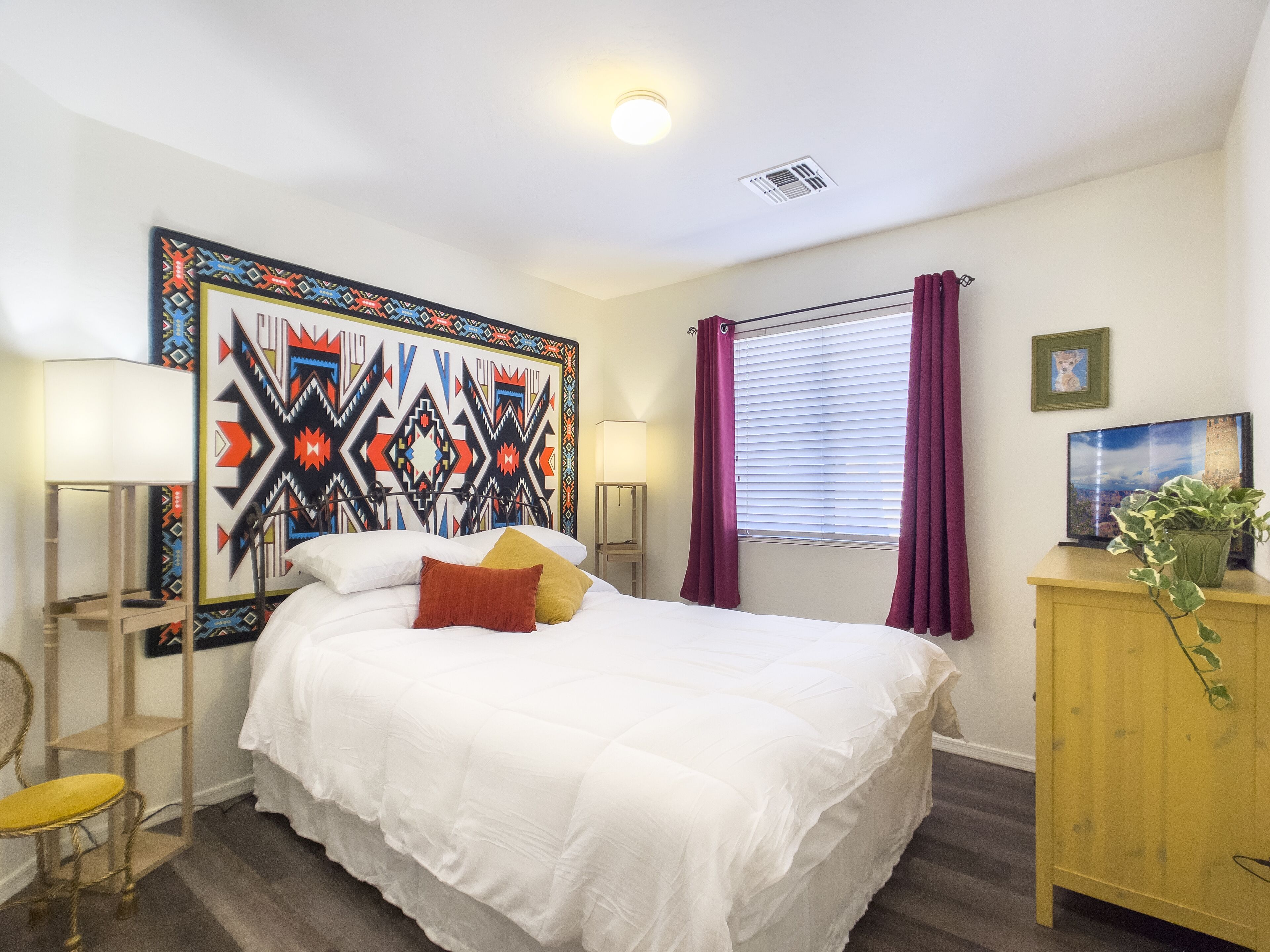 A neatly arranged bedroom with a large bed, a bedside table, a lamp, a television, and a decorative wall piece.