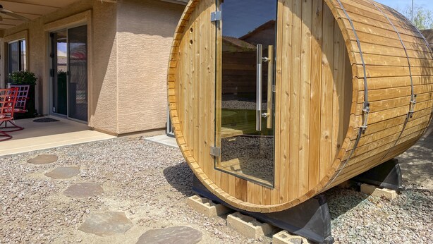 Un sauna en bois doté d’une porte coulissante, installé sur une terrasse en gravier, avec un bâtiment à toit de tuiles en arrière-plan.