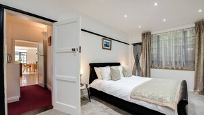 A bedroom with a large bed, a bedside table, a lamp, a window with curtains, and a door leading to another room.