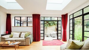 A spacious living room with a beige sofa, a wooden coffee table, and a red rug. Large windows with red curtains offer views of a garden and a brick building.