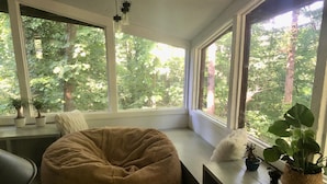 Un coin salon confortable avec un fauteuil en forme de poire et un banc, de grandes fenêtres donnant sur les arbres et des plantes en pot sur le rebord de la fenêtre.