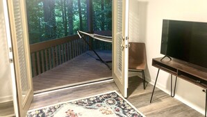 Une porte ouverte donnant sur un balcon doté d’un plancher en bois, d’un téléviseur posé sur un meuble et d’un tapis à motifs.